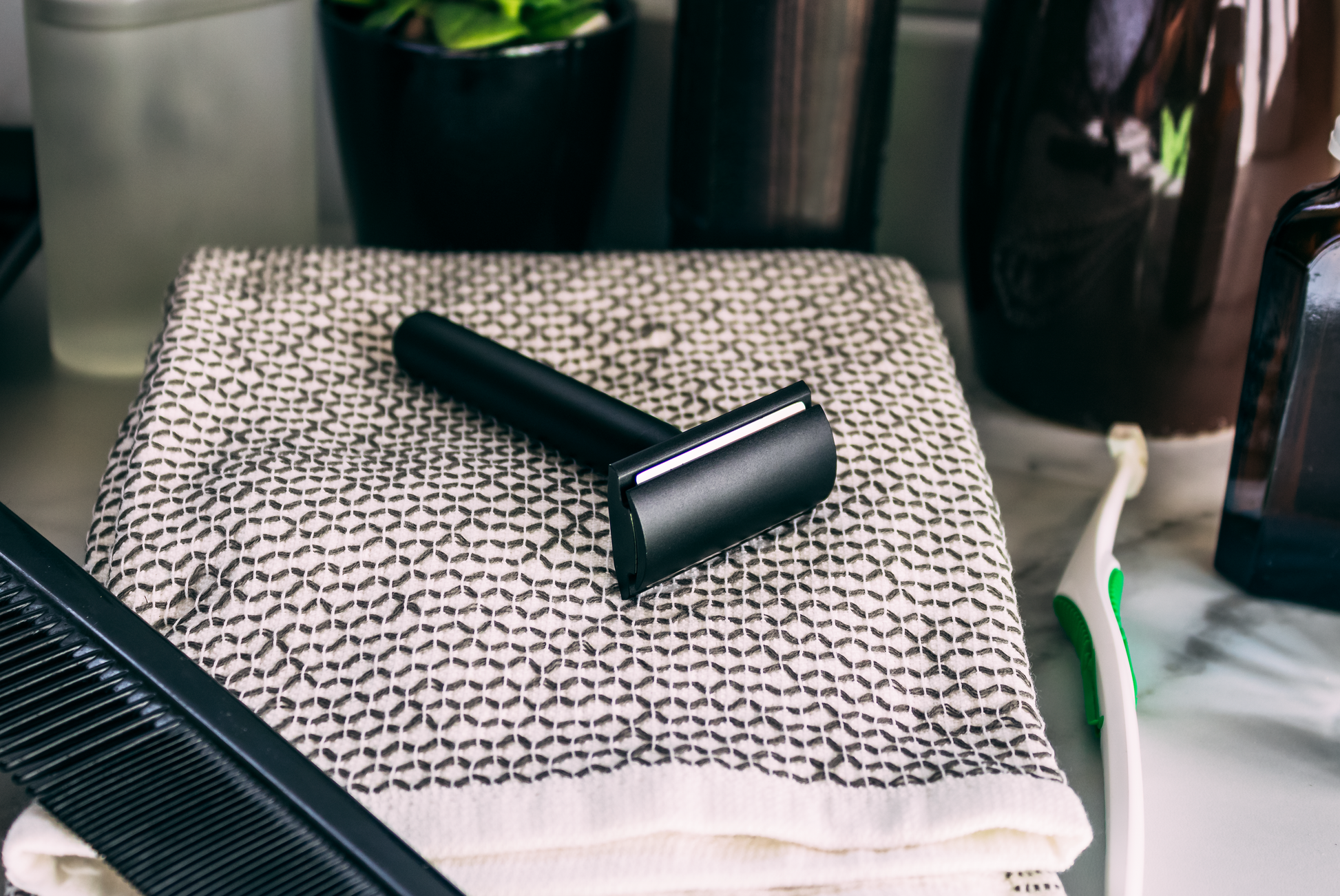 A Standard Razors Aluminum Series Double Edge Safety Razor (Deep Black) rests on a patterned towel, next to a black comb, electric toothbrush, and assorted containers in the background.