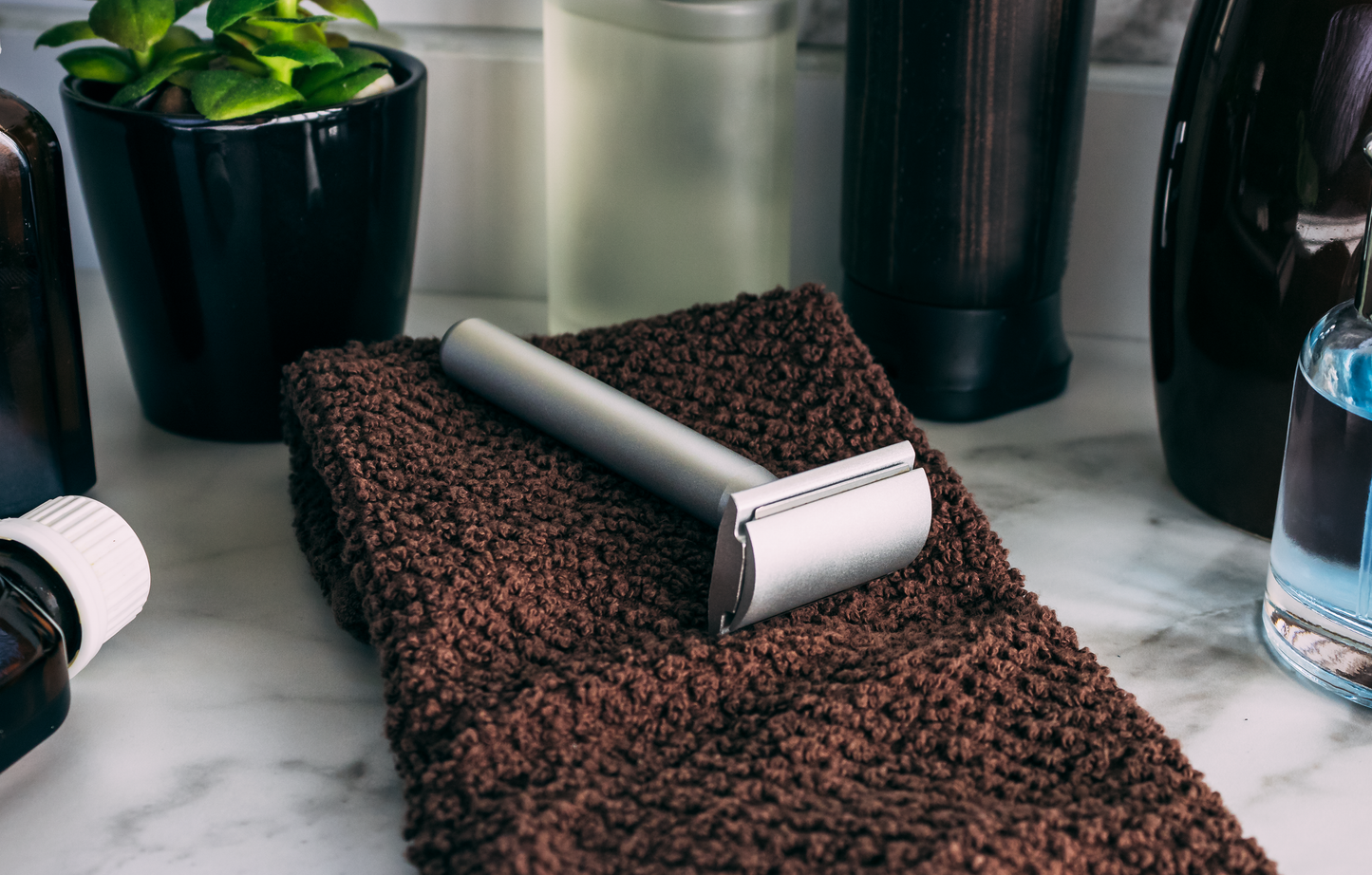 The Standard Razors Aluminum Series Double Edge Safety Razor (Raw Silver) by Standard Razors sits on a folded brown towel, with toiletry bottles and a small potted plant nearby on a marble countertop.