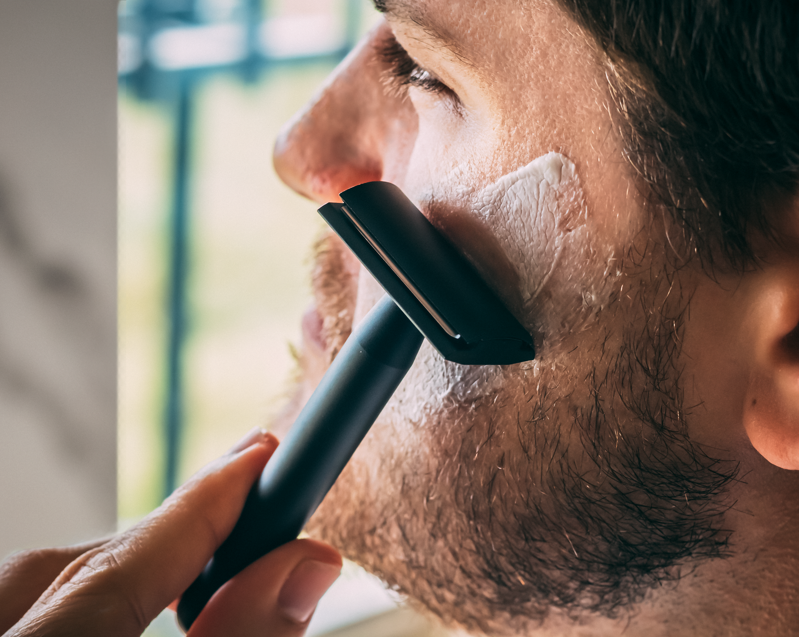 A close-up of a person shaving their bearded cheek with the Standard Razors Aluminum Double Edge Safety Razor — Deep Black, shaving cream applied to the skin.
