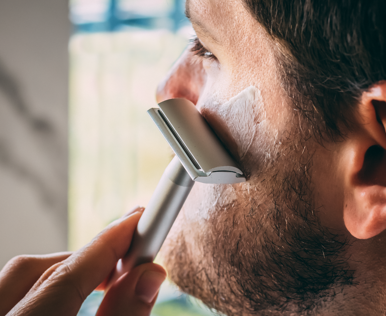 A close-up of a person shaving their cheek with the Aluminum Double Edge Safety Razor — Raw Silver by Standard Razors, applying shaving cream to their skin.